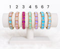 Pastel crystal stretch bracelets displayed on a white jewellery stand, featuring rectangular crystal stones in aqua, pink, lavender, blue, and iridescent tones with gold-tone accents.