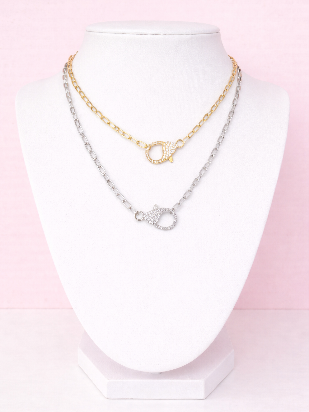 Two necklaces, one gold and one silver, on a mannequin head against a pink background.
