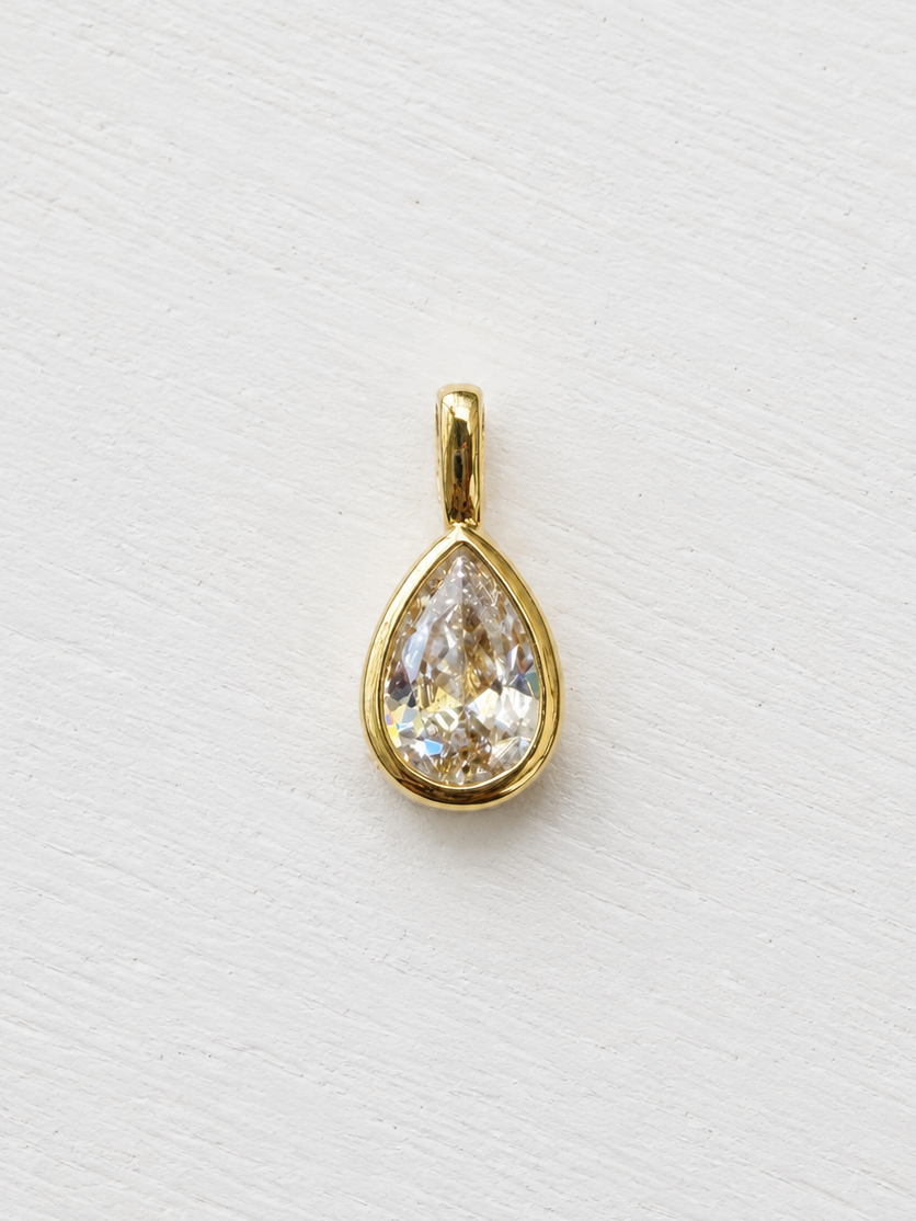 A gold teardrop pendant with a clear faceted gemstone, displayed on a light textured white background.
