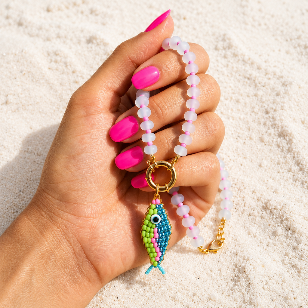 Hand holding the Lucky Catch beaded fish charm necklace with bright pink nails against white beach sand.