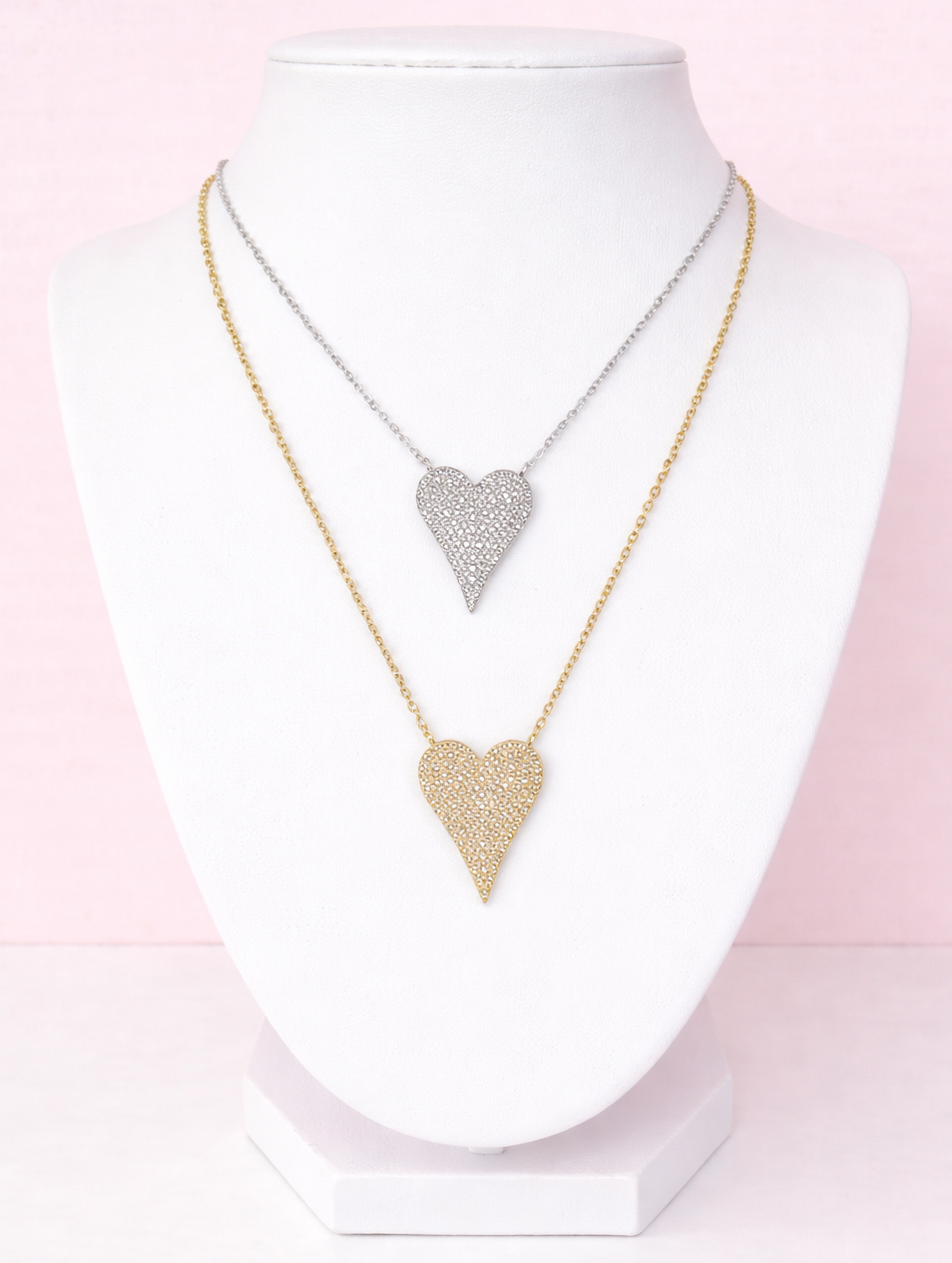 Two heart-shaped pavé necklaces on a mannequin with a pink gradient background