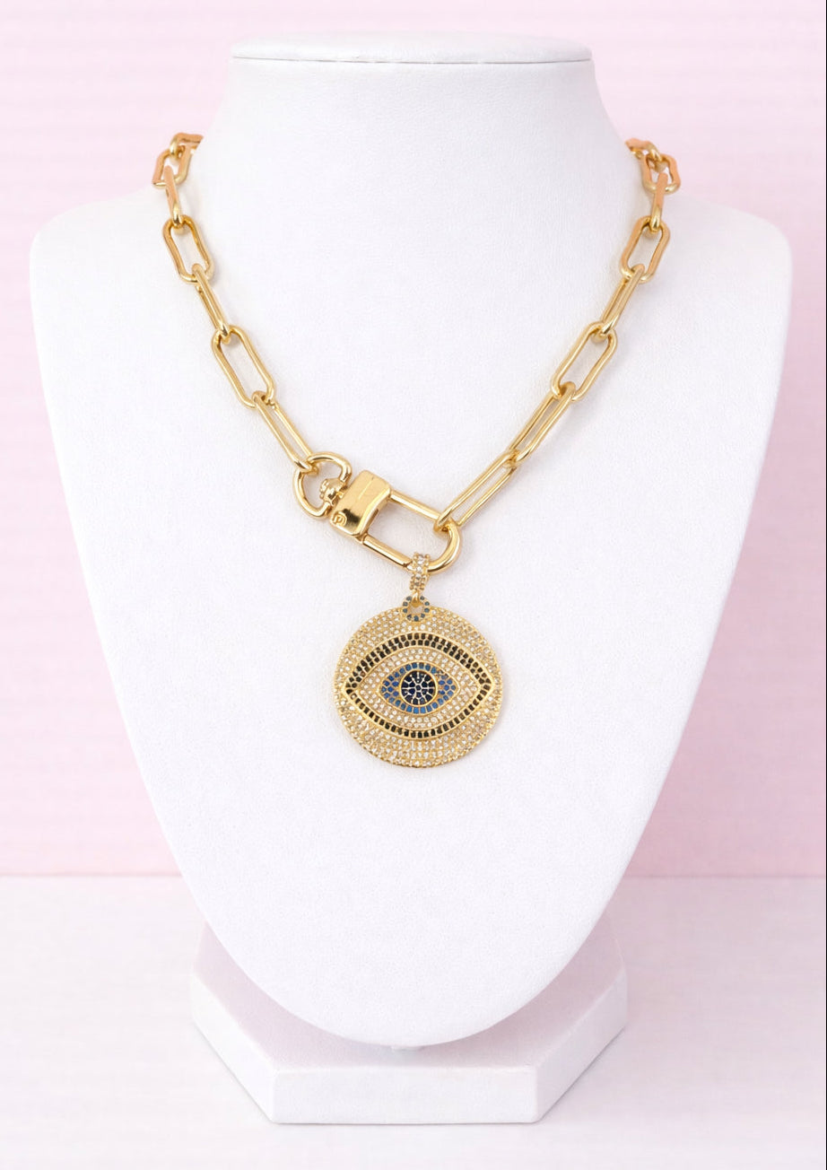 Gold necklace with a pavé evil eye pendant on a white stand against a pink background