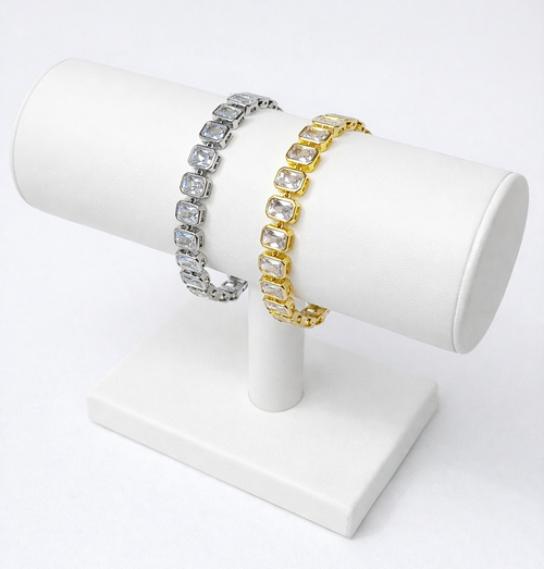 Two bracelets, one silver and one gold, on a white cylindrical stand against a white background.