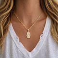 Gold necklace with a Hamsa hand pendant worn by a person in a white top.