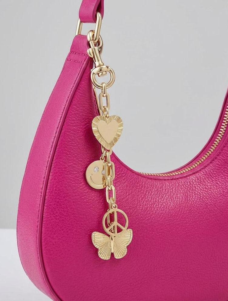 Gold tone, bag charm clipped to a pink hand bag featuring heart, smile face, peace symbol, and butterfly charms