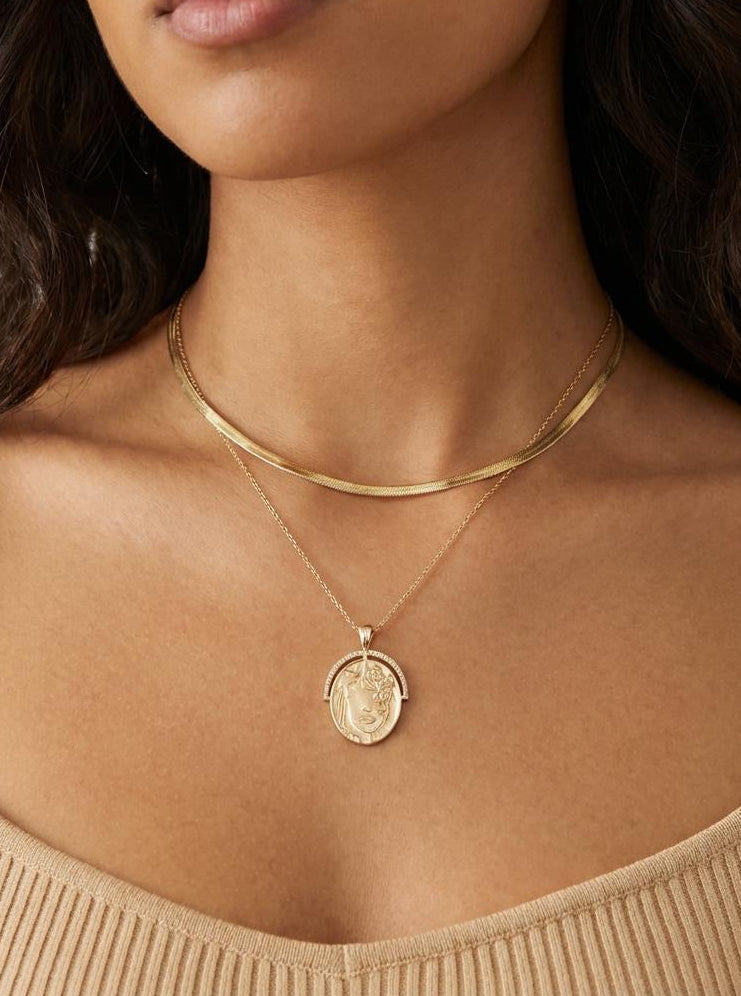 Model wearing a gold plated, oval pendant necklace with engraved detail layered