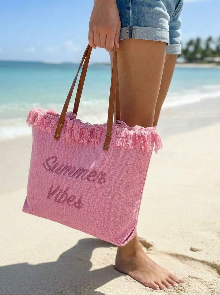 Person holding a pink beach bag with 'Summer Vibes' text on a sandy beach.