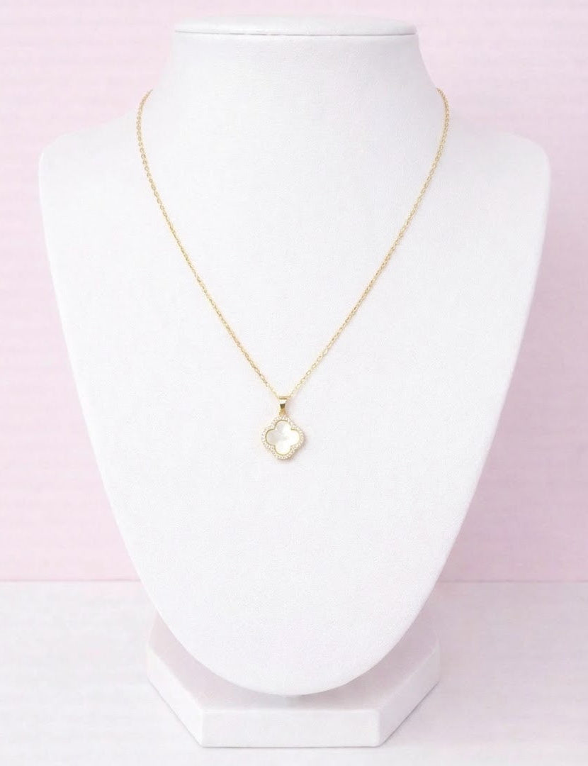Gold clover necklace with a heart pendant on a white stand against a pink gradient background